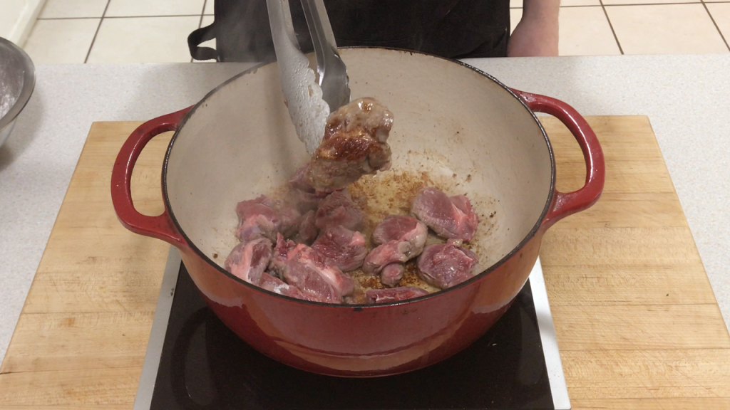 The lamb is being browned in batches in a red dutch oven.